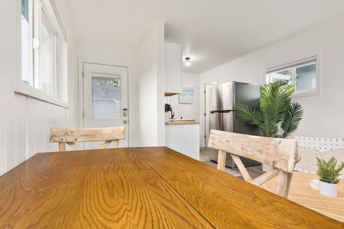 A cozy open-plan area featuring a wooden dining table set close to a bright kitchen with contemporary appliances. Fresh greenery adds a touch of nature, while a doorway and ample natural light suggest seamless flow between spaces.
