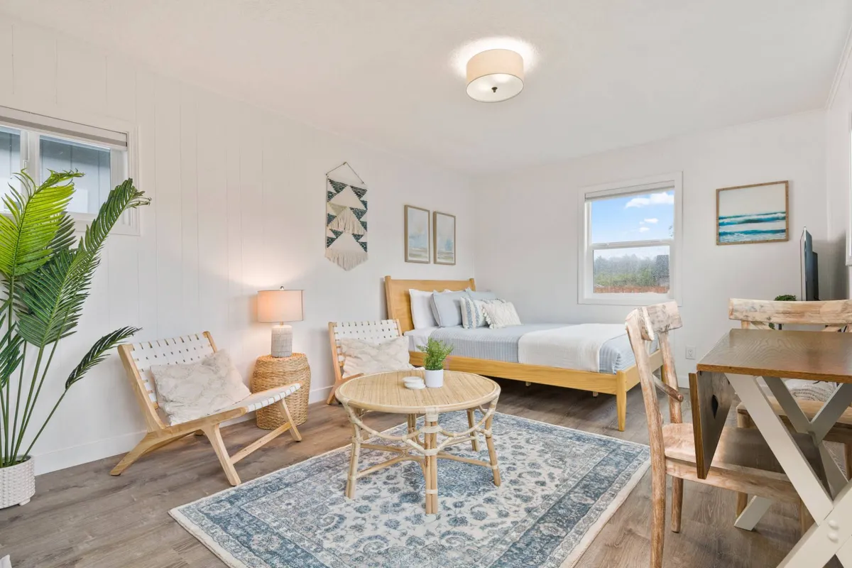 A restful bedroom scene with a bed dressed in light blue linens, a round coffee table, decorative wall art, and a sunlit window. The space is tranquil, comfortable, and thoughtfully arranged for relaxation throughout the day.