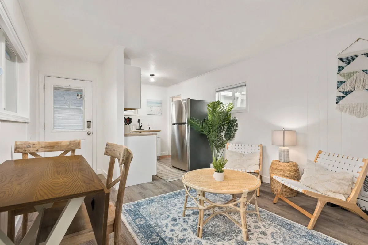 The photo showcases a sunlit open-plan living area featuring a cozy rattan coffee table, a leafy potted plant, inviting wooden dining table, plush chairs, and a well-equipped kitchen—perfect for intimate gatherings and relaxed coastal living.