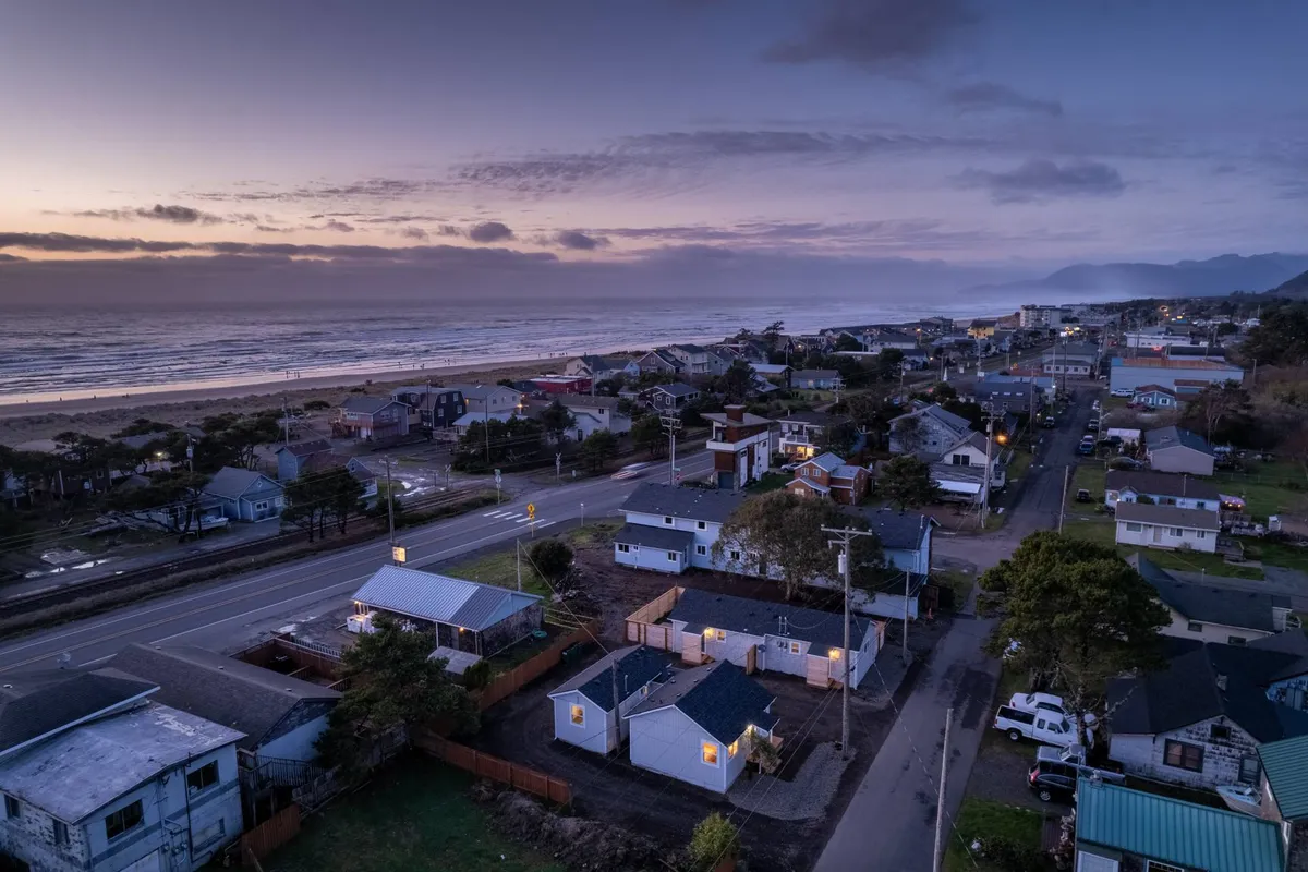 A peaceful streetscape where houses are nestled close to the ocean, with leafy trees and open skies. The beach and town are both within sight, hinting at the convenience of exploring local shops, cafes, and restaurants by foot.