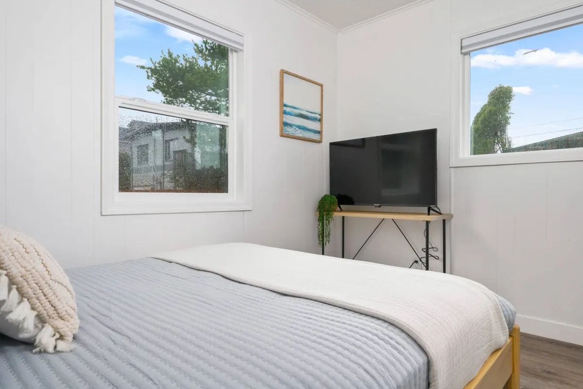 A serene bedroom with gray bedding, a television on a stand, ocean-themed wall art, and large windows showcasing surrounding greenery. The space is tidy and inviting, filled with natural light.