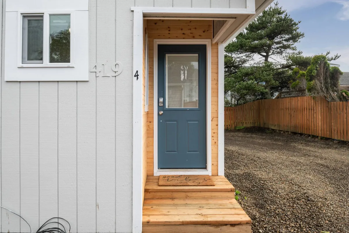 A close view of a cottage’s blue front door, wooden steps, and a welcoming mat, surrounded by lush greenery. The porch area looks ideal for relaxing or interacting casually with neighbors as you come and go.