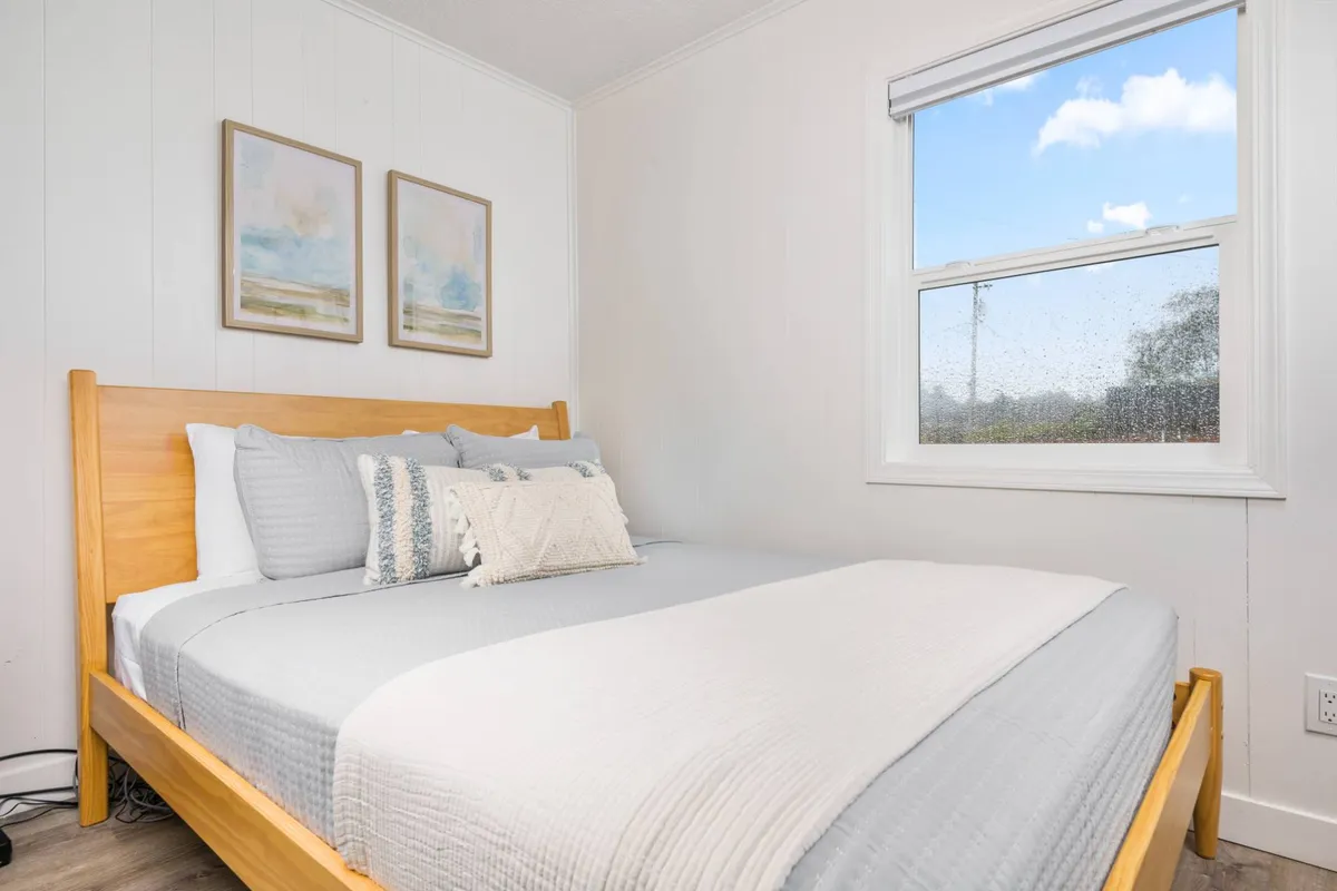 A tranquil bedroom space featuring a warm wooden bed frame adorned with decorative pillows. Framed artwork hangs above the bed, and a large window lets in abundant natural light, softly illuminating the room. The ambiance is serene and inviting, underscoring relaxation and comfort.
