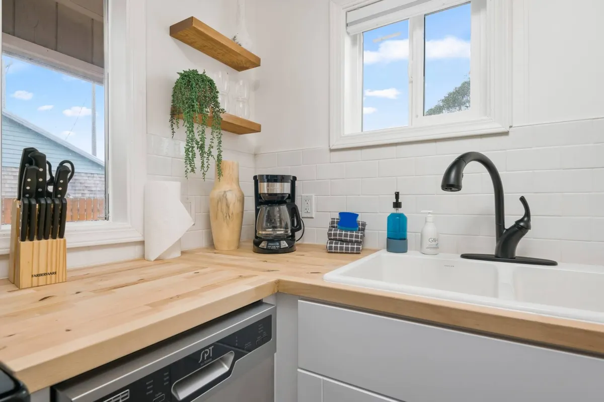 A charming kitchen corner featuring a wooden countertop, dishwasher, coffee maker, and open shelves with decorative objects. A window offers a pleasant view, letting in natural light and connecting the indoors to the lively environment outside.
